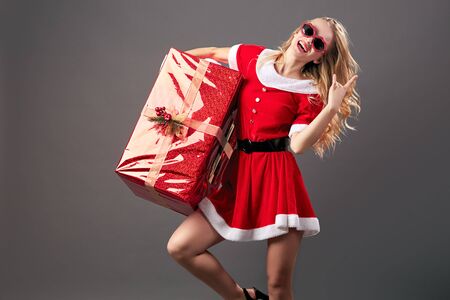 Young and beautiful mrs.Santa Claus in sunglasses dressed in the red robe, white gloves and high heels holds the huge  Christmas present on the gray background.の写真素材