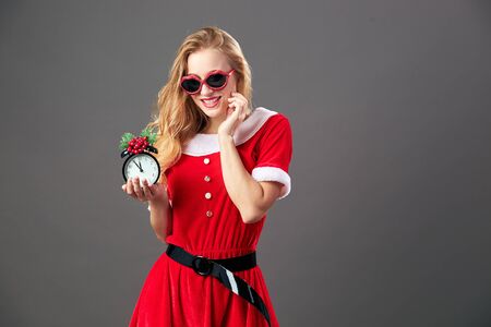 Young and beautiful mrs.Santa Claus in sunglasses dressed in the red robe and white gloves is holding a clock that shows five to twelve. Christmas Eveの写真素材