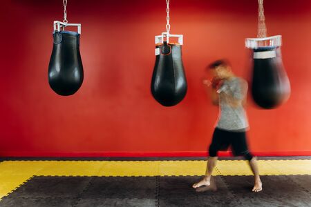 Guy dressed in the grey t-shirt and black shorts works out with a black punching bag against a red wall in the gymの写真素材