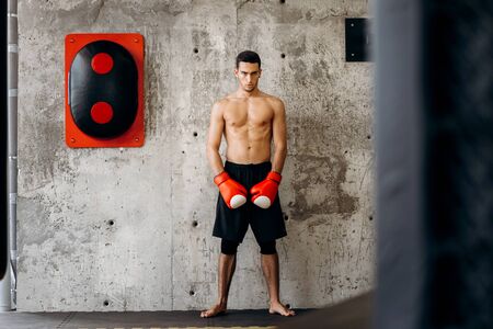 Brutal dark-haired guy with a naked torso and the red boxing gloves stands against a concrete wall next to equipment for boxingの写真素材