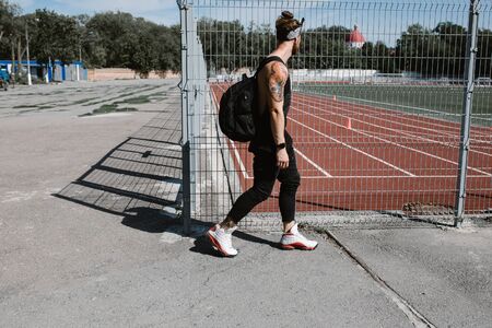 Athletic guy in headband dressed in black sport clothes with backpack on his shoulders walks along...