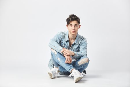 Dark-haired guy dressed in a white t-shirt, jeans and a denim jacket is sitting cross-legged on the floor on the white background in the studioの写真素材