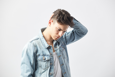 Thoughtful dark-haired guy dressed in a white t-shirt and a denim jacket holds his hand on the head  on the white background in the studioの写真素材