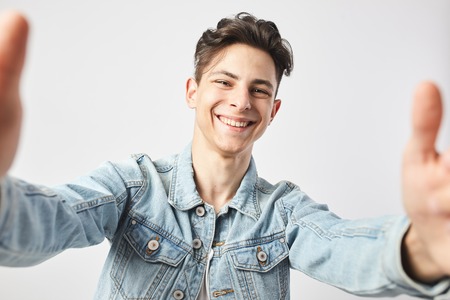 Smiling dark-haired guy dressed in a white t-shirt and a denim jacket  makes a salfie on the white...