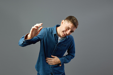 Laughing guy dressed in a jeans shirt keeps his hand on the stomach in the studio on the gray backgroundの写真素材