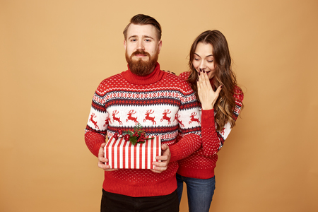 Guy dressed in red and white sweater with deer holds a Christmas present in his hands and a girl looks out from behind his back on a beige background in the studioの写真素材
