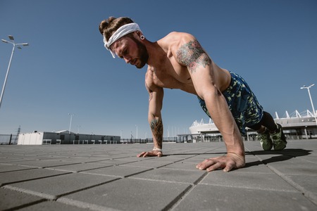 Handsome shirtless sportsman looking away doing push up exercise on workout ground outdoors at...