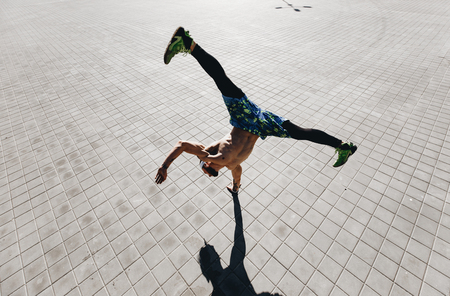 Young athletic man with a naked torso with tattoos dressed in the  black leggings and blue shorts doing the trick standing on one hand on the paving slabs in the streetの写真素材