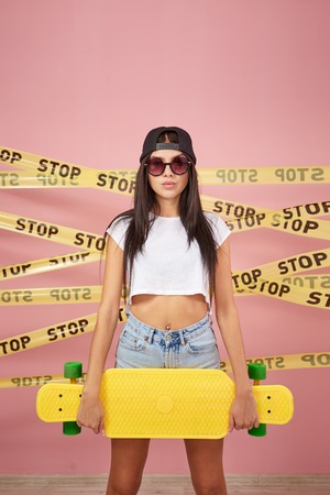 Young dark-haired girl in pink sunglasses and cap dressed in jeans shorts and white top stands with...