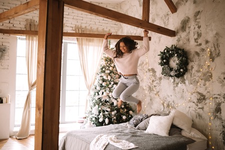 Charming girl dressed in white sweater and pants jumps on the bed with gray blanket and white pillows in a cozy decorated room with a New Year treeの写真素材