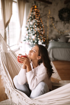 Charming dark-haired girl dressed in beige sweater and pants holds a red cup sitting in a hammock in a cozy decorated room with a New Year treeの写真素材