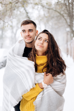 Charming dark-haired girl dressed in a yellow sweater and her boyfriend wrapped together in a gray scarf are standing in a snowy street on a winter dayの写真素材