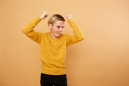 Joyful young blond guy dressed in yellow sweater is posing in the studio on the beige backgroundの写真素材