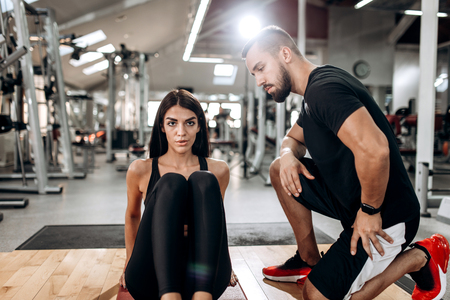 Slim girl dressed in black sports top and tights is doing exercises for the abdominals  on the fitness mat in the gym next to the  coachの写真素材