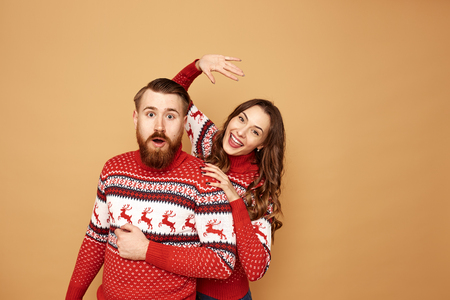 Funny girl and a guy dressed in red and white sweaters with deer stand together on a beige background in the studioの写真素材