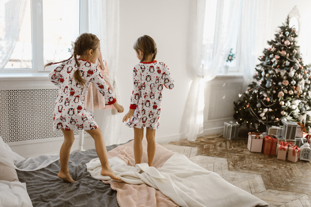 Two charming little girls in their pajamas are having fun jumping on a bed in a sunlit cozy bedroom with New Years tree and giftsの写真素材