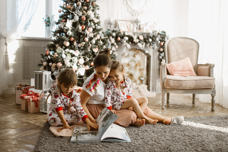Young beautiful mother with two little daughters sit on the carpet and read book near the New Years tree with gifts in the light cozy room with armchair and fireplaceの写真素材