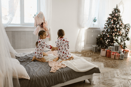 Two charming little girls in their pajamas are having fun jumping on a bed in a sunlit cozy bedroom with New Years treeの写真素材
