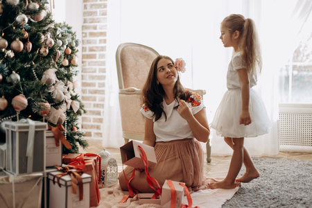 Happy young mother with flower in her hair and her  little daughter in nice dress sit near the New Years tree and open  New Years gifts in the light cozy roomの写真素材