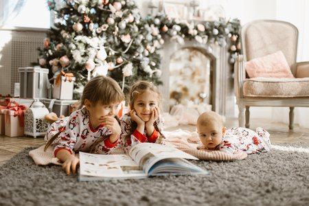 Two little sisters and a tiny brother lie on the carpet and read book near the New Years tree with gifts in the light cozy room with armchair and fireplaceの写真素材
