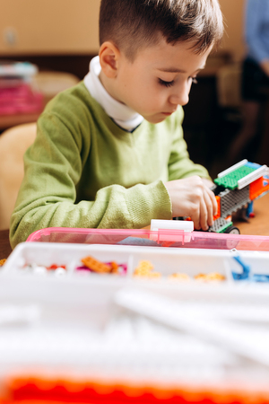 Focused boy  dressed in green sweater sits at the table in the robotics school and makes a robot...