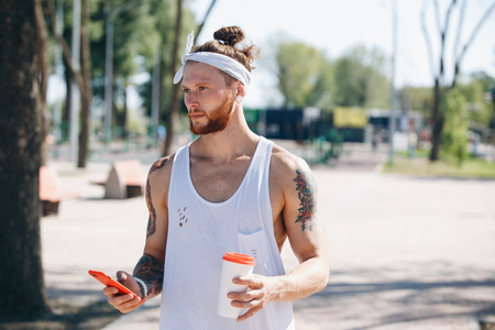 Young man with white headband dressed in a white t-shirt using mobile phone and holds a plastic cup in his hand on the sports ground outside on a sunny dayの写真素材