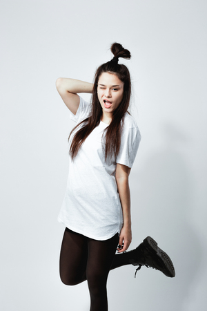 Young dark-haired girl with a funny hairstyle dressed in white t-shirt, black tights and shoes winks posing on the white background in the studioの写真素材