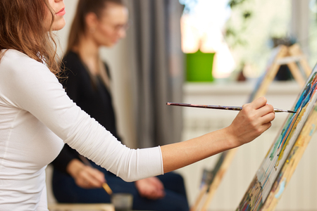 Girl with brown curly hair dressed in white blouse paints a picture at the easel in the drawing schoolの写真素材