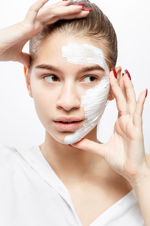 Young woman dressed in white clothes with a white cosmetic mask on a half of her face holds her hands at her face on the white backgroundの写真素材