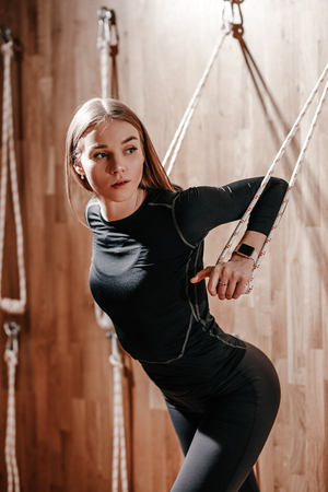 Slim young girl dressed in a black sportswear is doing stretching on special rope equipment in the modern gym with wooden decorationの写真素材