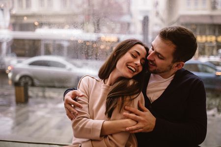 Loving guy hugs his beautiful happy girlfriend sitting on the windowsill in a cozy cafe.の写真素材
