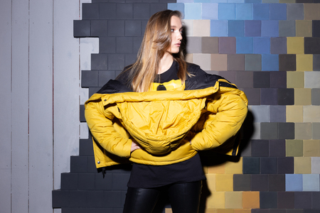Portrait of a young fashionable girl in black urban outfit and yellow jacket standing at night on the street opposite the wall.の写真素材
