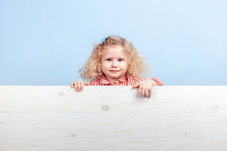 Funny little curly girl in a striped red and white dress and blue flower on her hair stands behind the wooden board against a blue wallの写真素材
