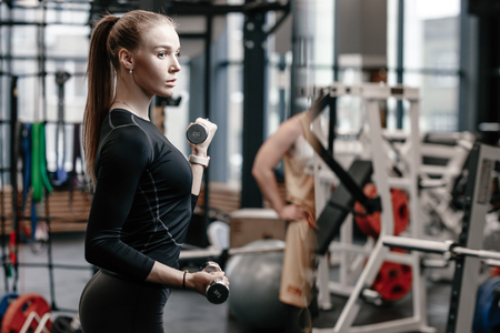 Slender young girl dressed in a black sportswear is doing exercises with dumbbells in the modern gym with lots of sport equipmentの写真素材