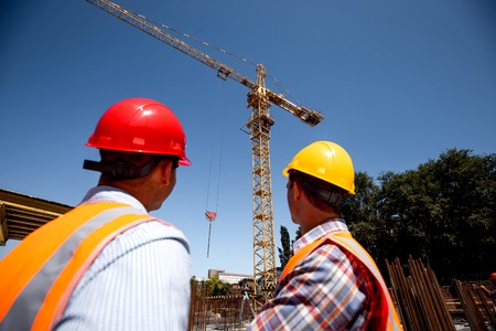 Two men dressed in orange work vests and  helmets look at th crane on the building site