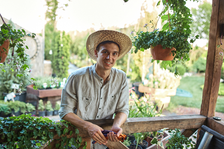 Guy gardener in a straw hat is standing next to a wooden veranda with a lot of plants on a sunny day.の写真素材