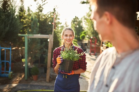 Beautiful smiling girl gardener holds pot with plants and looking at the guy in the wonderful garden on a sunny dayの写真素材