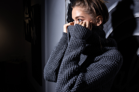 Portrait of a young fashionable girl in a gray sweater standing at night on the street opposite the wall. The mouth and nose of the girl is covered with a sweater.の写真素材