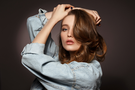 Beautiful brunette girl with long flowing hair dressed in jeans jacket poses holding her hands on her head on the dark background in the studioの写真素材