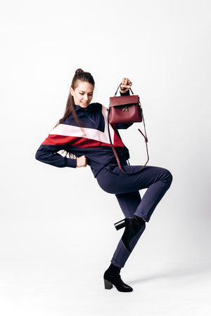 Girl dressed in sporty blue suit with a red and white print on a sweatshirt and heels poses with bag in her hands on the white background in the studioの写真素材