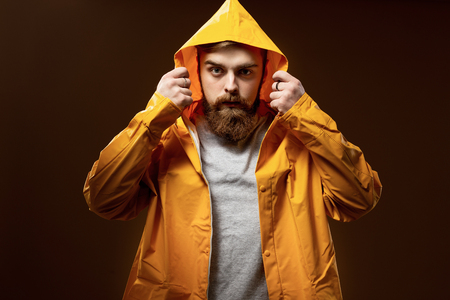 Red-haired guy with a beard and mustache dressed in a gray t-shirt and yellow jacket with a hood is posing on a brown background in the studioの写真素材
