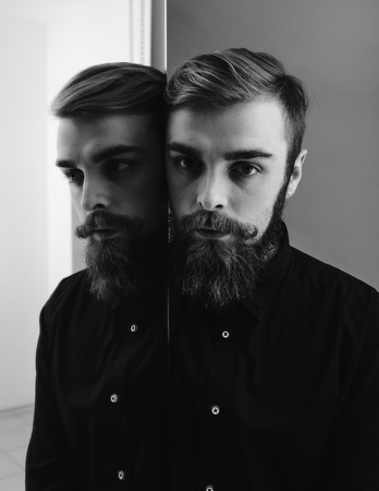 Black and white photo of a man with a beard and stylish hairdo dressed in the black shirt standing next to the mirror with his reflectionの写真素材