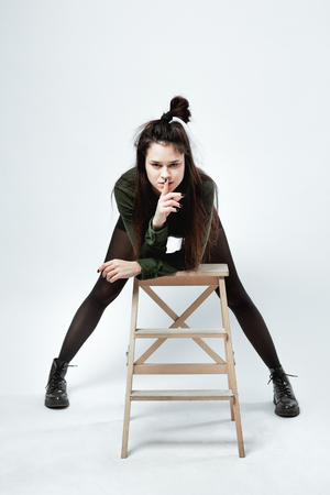 Young stylish dark-haired girl with a funny hairstyle dressed in green shirt, black shorts , tights and shoes is posing with  the wooden stool on the white background in the studioの写真素材