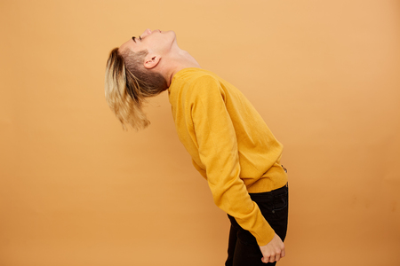 Stylish blond guy dressed in yellow sweater and black jeans is posing on the beige background in the studioの写真素材
