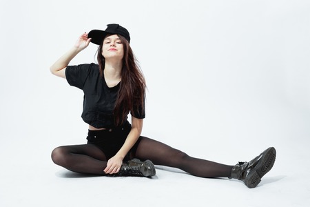 Funny young stylish girl dressed in a black top, shorts, tights and cap  sits on the floor  on the white background in the studioの写真素材