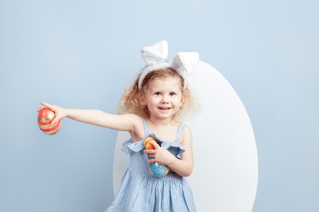 Charming curly girl in the light-blue dress with bunny ears on her head holds the dyed eggs in her hands on the background of a big white egg on a blue wall. Easter bunnyの写真素材