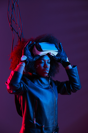 Curly dark haired girl dressed in a black leather jacket and gloves is wearing  the virtual reality glasses on her head in the dark studio with neon lightの写真素材