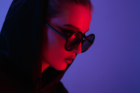 Portrait of fashion young girl in a black sweater with a hood and round sunglasses in red and blue neon light in the studioの写真素材