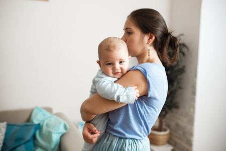 Young mother dressed in light blue t-shirt and skirt is holding her tiny son on her arms in the room at homeの写真素材