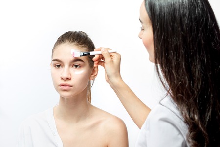 Cosmetologist applies with a brush white cosmetic mask on the face of young brown-haired girl